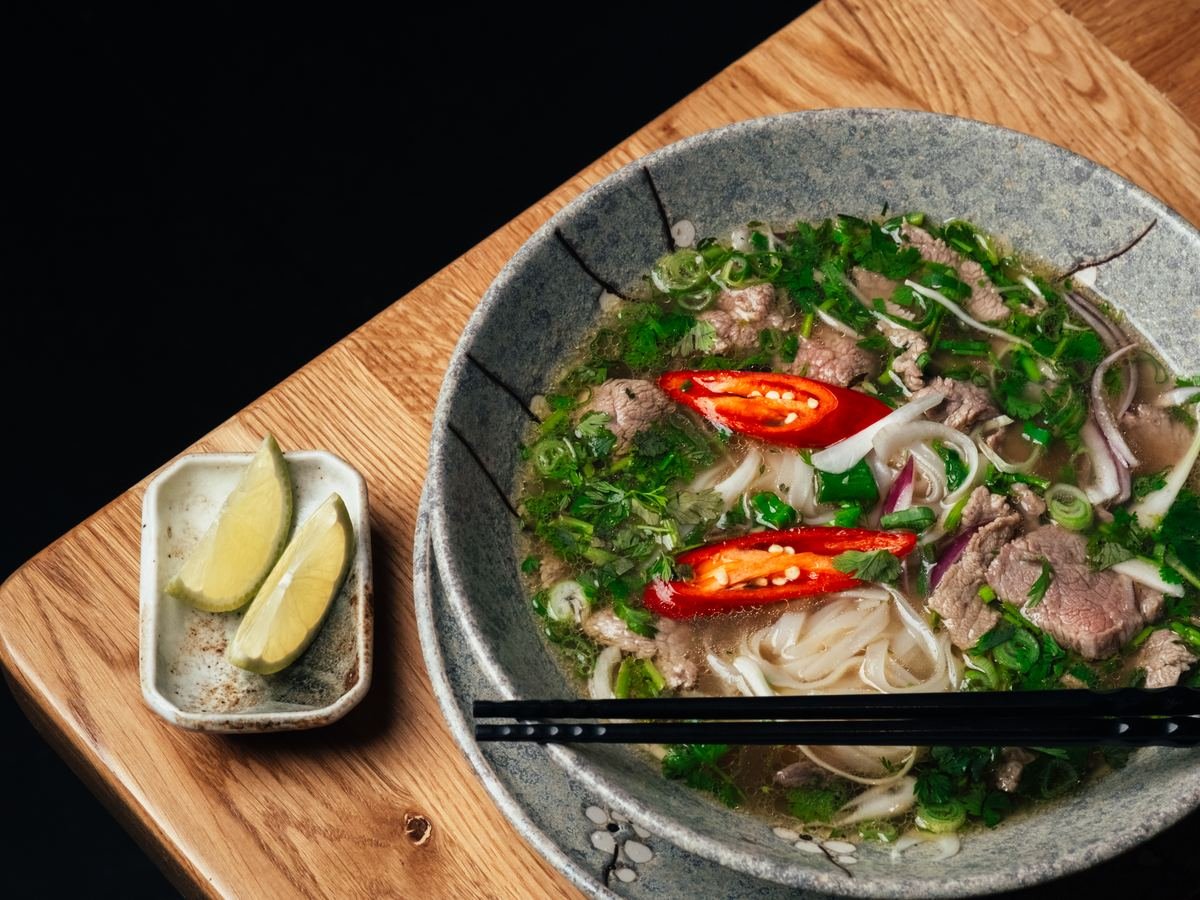 Schale Phở mit Rind, Reisbandnudeln, frischen Kräutern und Chili, daneben Limetten
